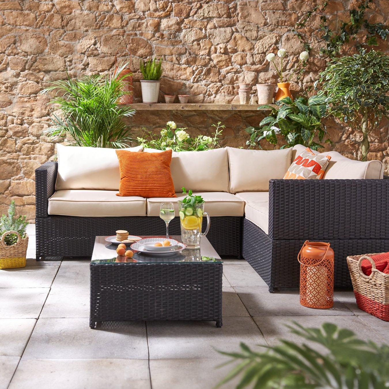 Black patio with a wicker sofa, cream cushions, and orange pillows. A glass-top table holds drinks and snacks. Surrounding plants add a lush touch.