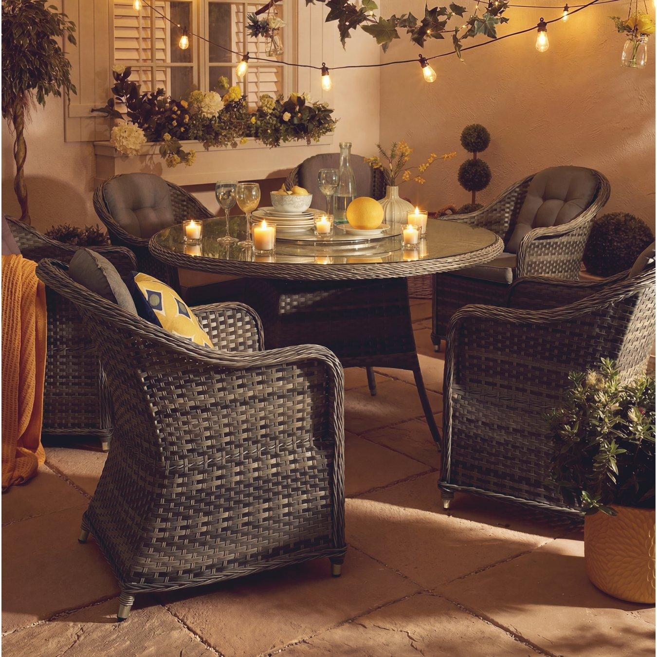 Cosy patio scene with Poly-rattan garden furniture set of chairs and a round table set for dining. String lights and candles create a warm, inviting glow. 