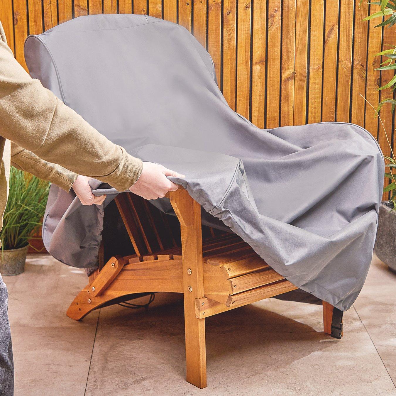 Person removing a grey cover from a wooden patio chair on a tiled outdoor patio with a wooden fence and potted plants in the background.