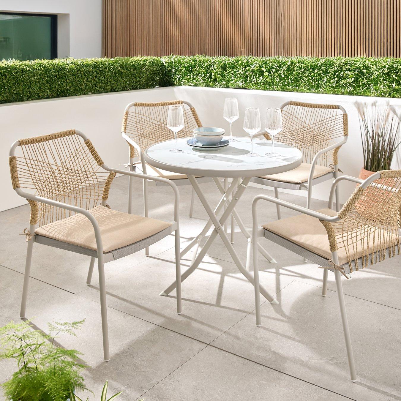 Cosy outdoor patio with a white round table, four wicker chairs, and elegant glassware. Surrounded by green hedge, sunny and inviting.