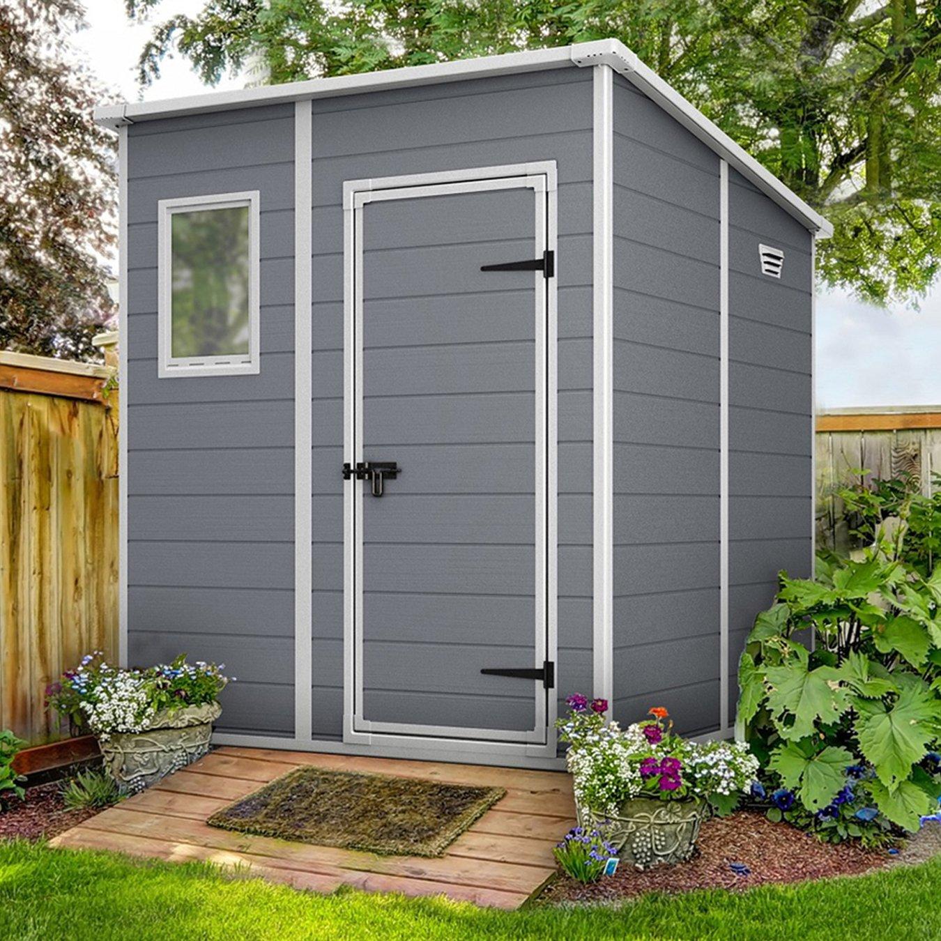 A grey garden shed with a white-trimmed door and window sits on a wooden deck, surrounded by blooming flowers and lush greenery.