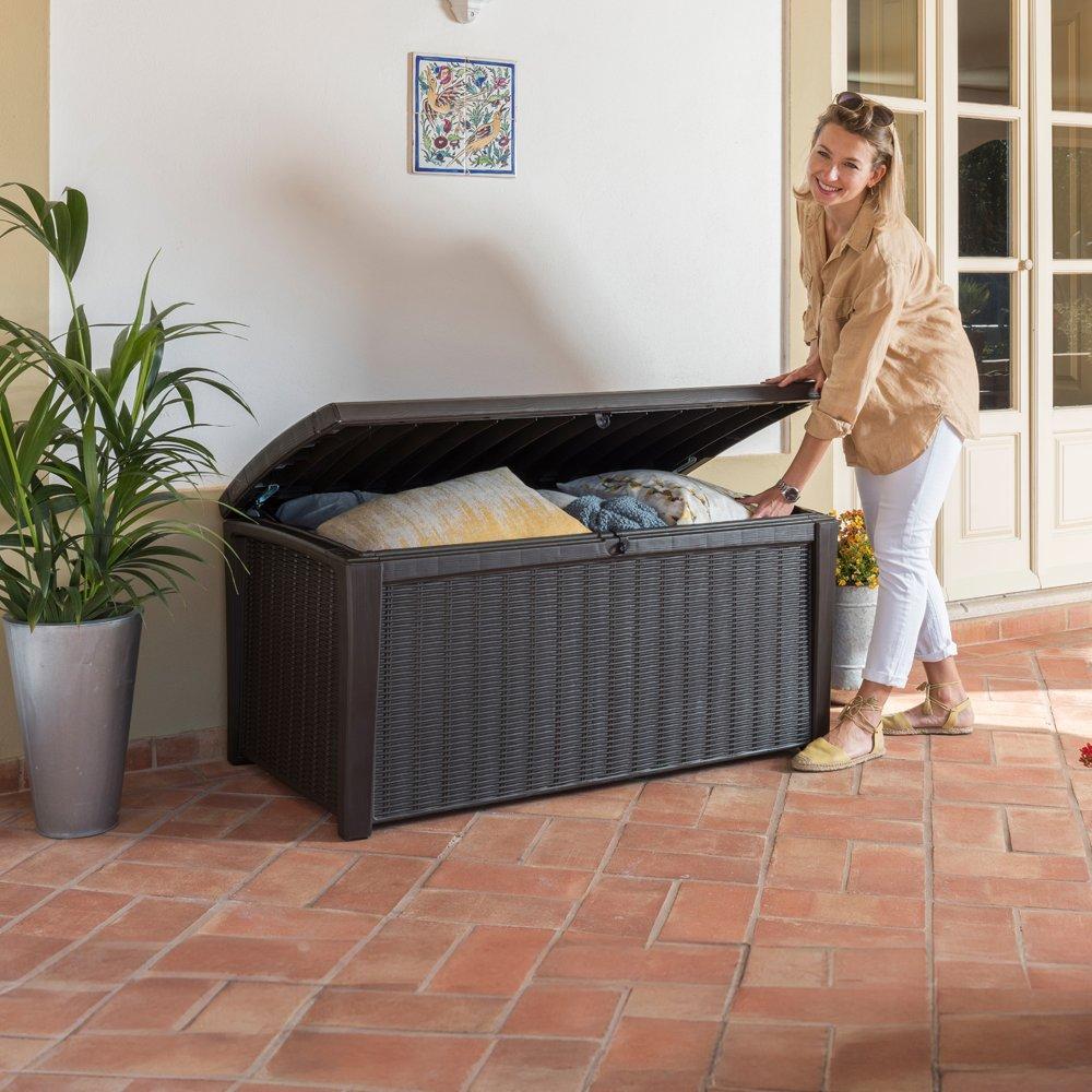 A woman in casual clothing smiles while opening a large, garden storage box on a terracotta-tiled patio, with cushions inside and a potted plant nearby.