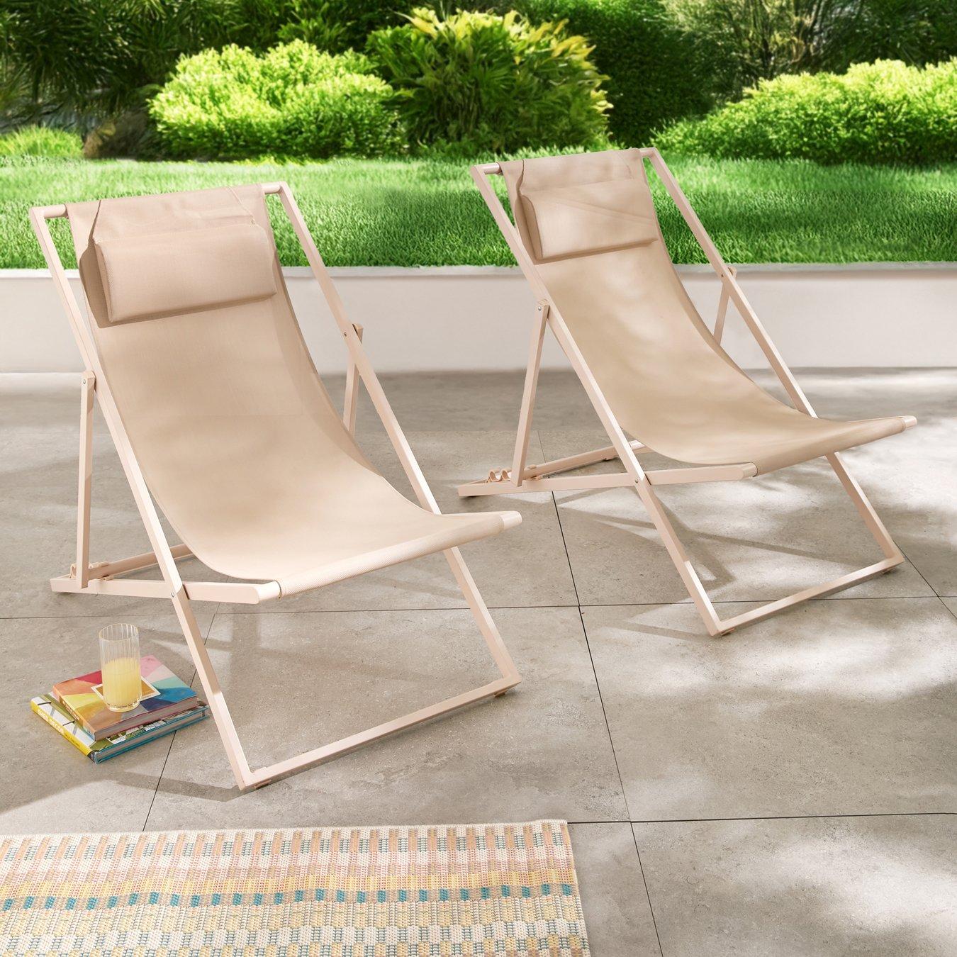 Two beige sling sun loungers sit on a tiled patio, facing away from a lush green lawn. A colourful rug and a glass of juice on books add a relaxed, summer vibe.