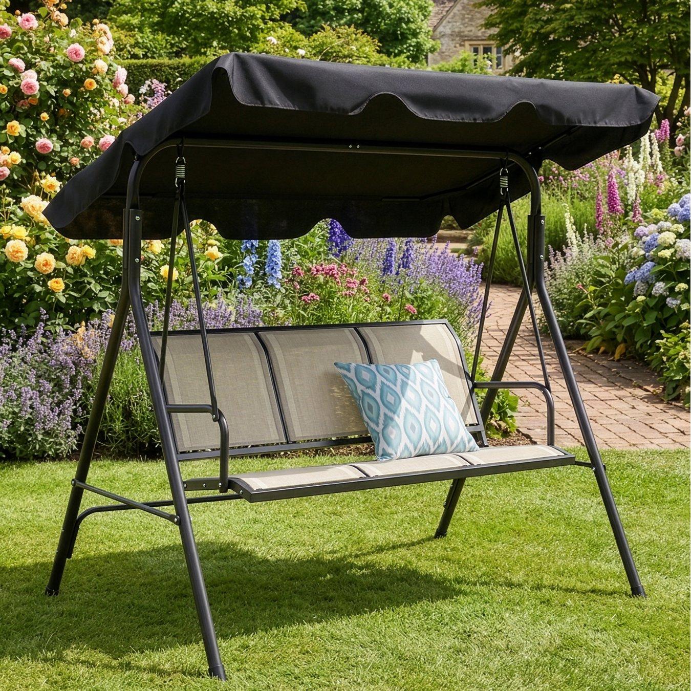 A black canopy garden swing set sits on green grass, surrounded by colourful flowers and a brick path, creating a serene garden scene.