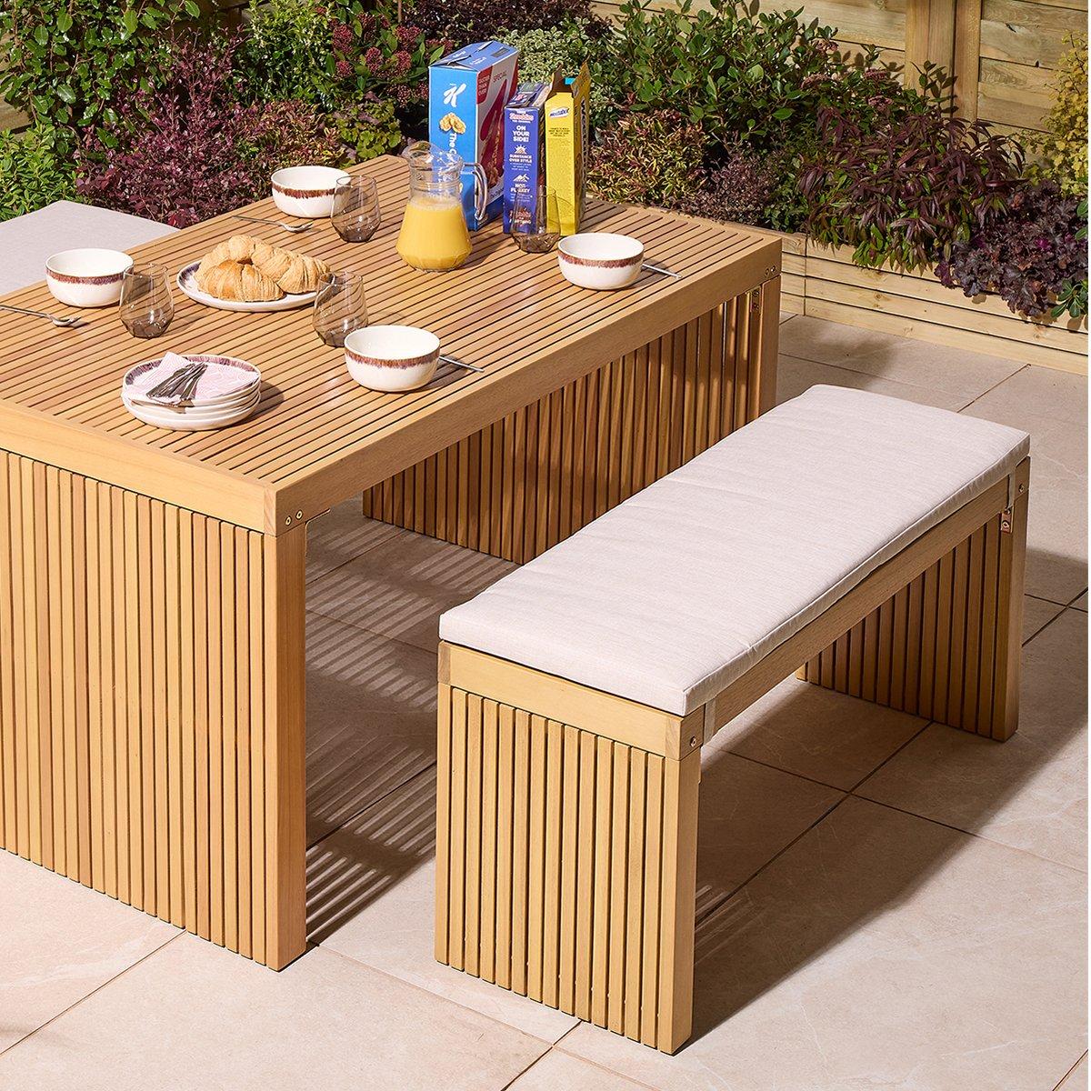 Wooden patio table set for breakfast outdoors. Table holds bowls, glasses, croissants, cereal boxes, and orange juice, surrounded by lush greenery.