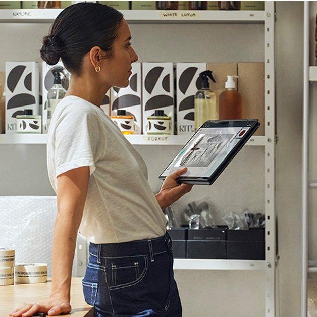 Woman in retail shop stands looking at products while holding a 2-in-1 laptop as a tablet.