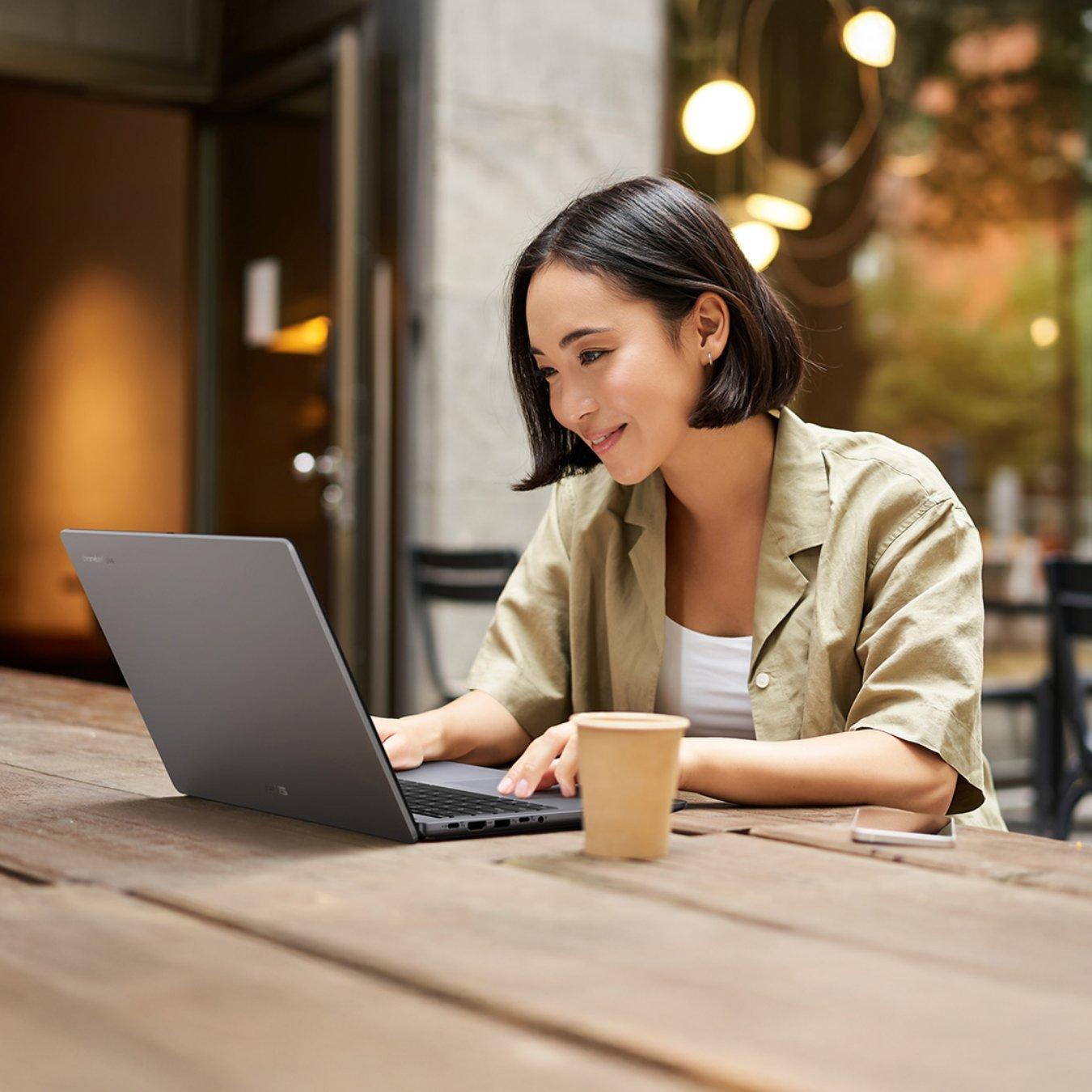 Woman wearing a green shirt sits at a wooden table in a coffee shop. On top of the table is an open Chromebook laptop.