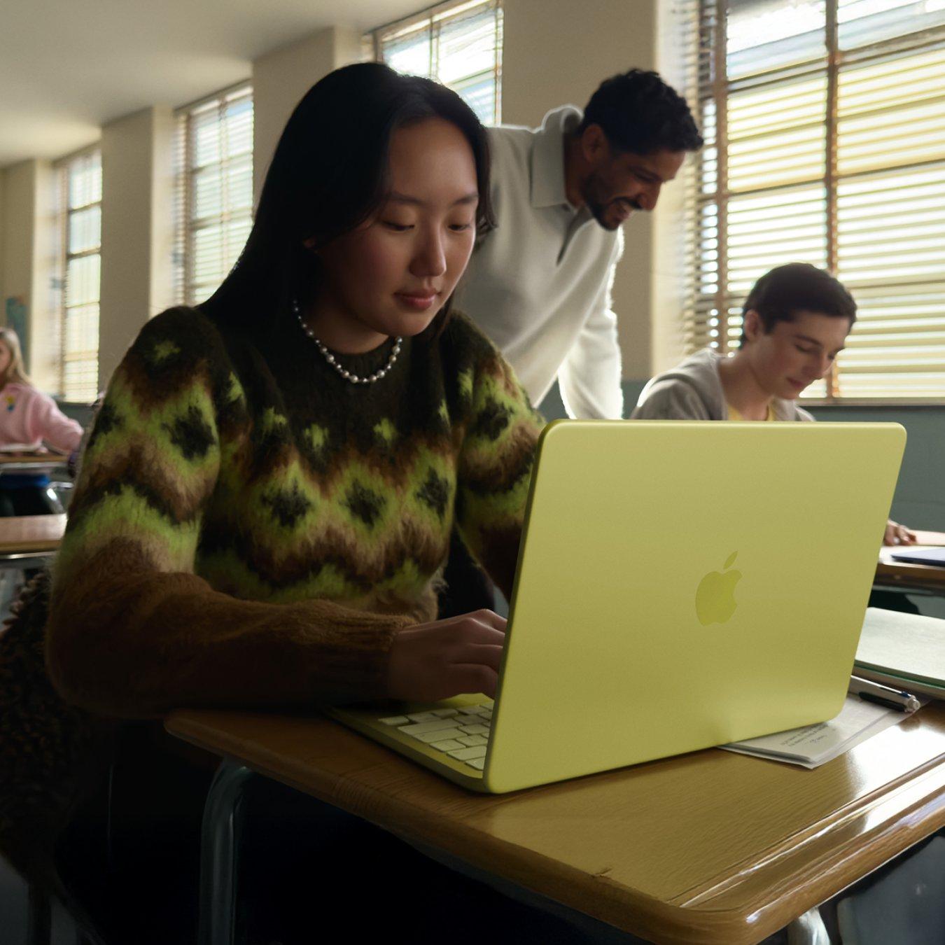 Student sits at her wooden desk in a classroom looking at work on her neon yellow MacBook Neo laptop.