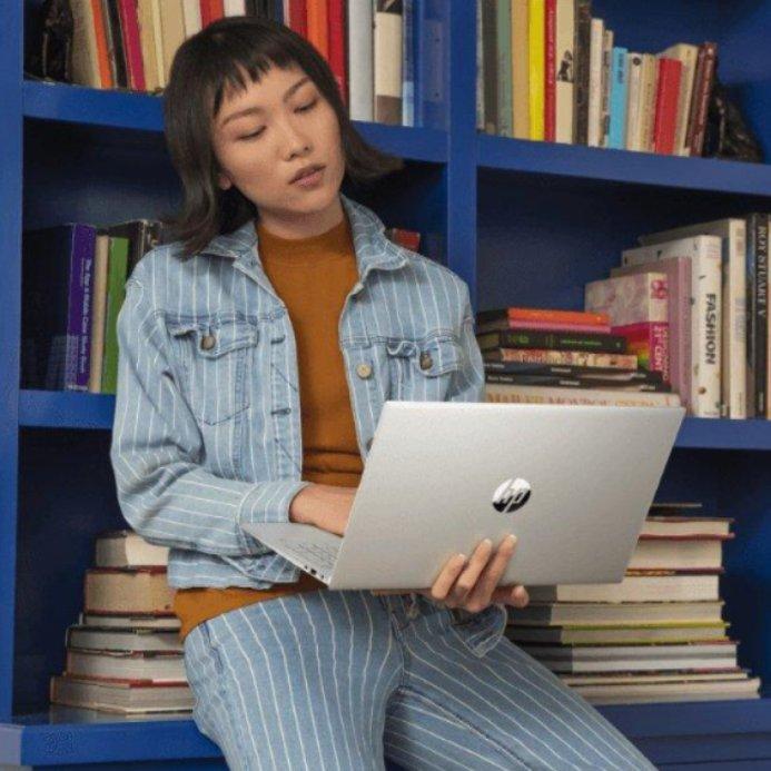 Woman in a denim co-ord leans on a blue bookcase while holding a silver HP laptop.