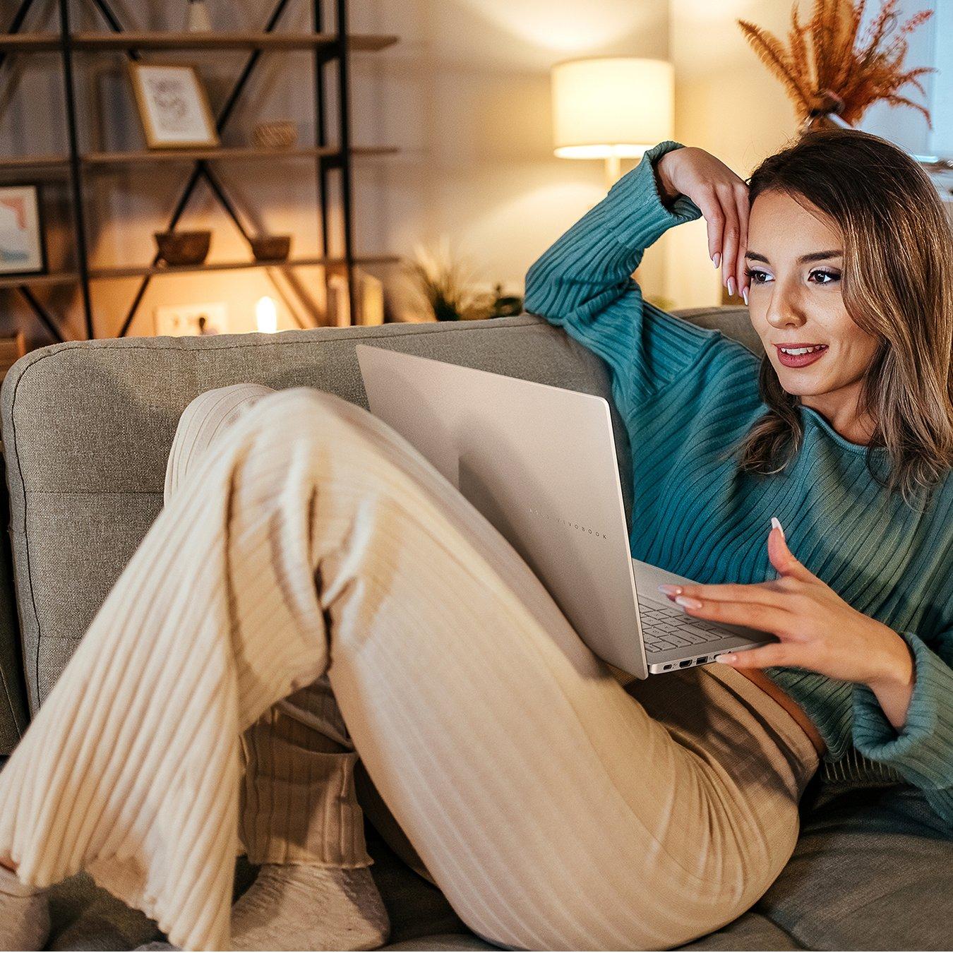 Woman relaxed on the sofa, wearing cream joggers and a teal jumper. On her lap rests a silver Windows laptop.