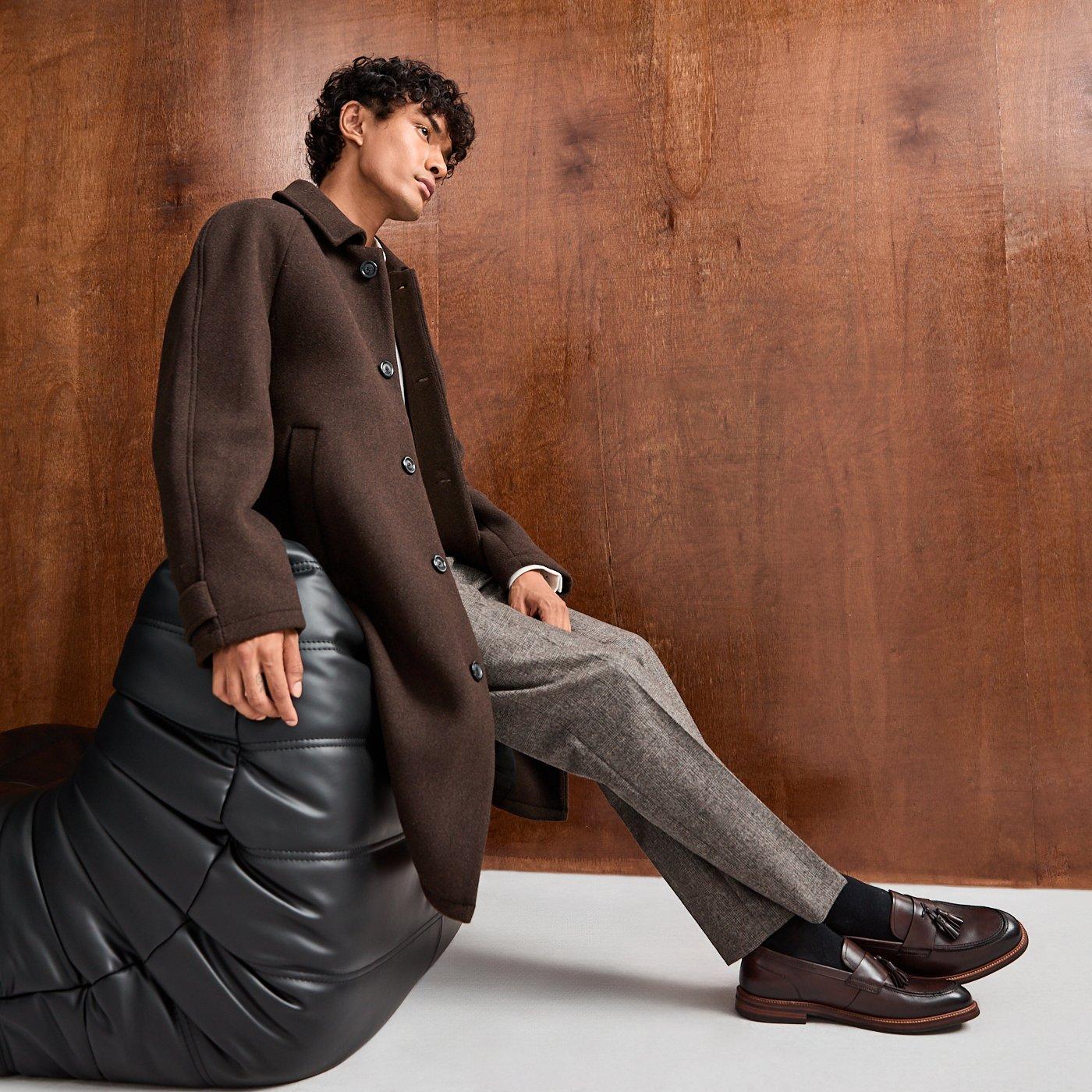 Man in a smart casual outfit consisting of an overcoat, trousers and formal shoes leaning against a black sofa.