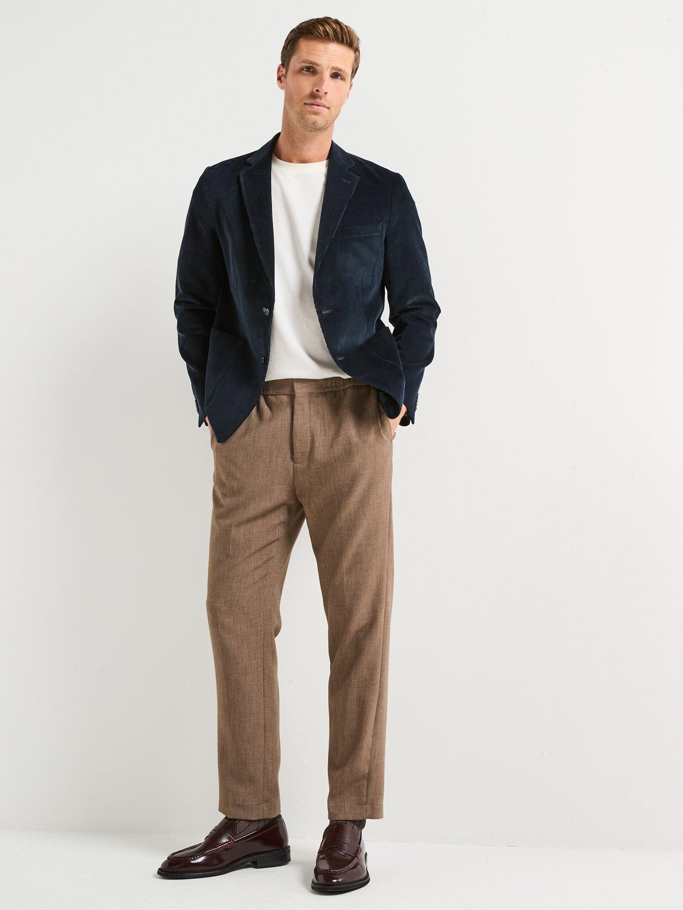 Man models while wearing the perfect smart causal outfit. The outfit consists of a plain white t-shirt tucked into brown cord trousers with brown boots and a navy velvet blazer.