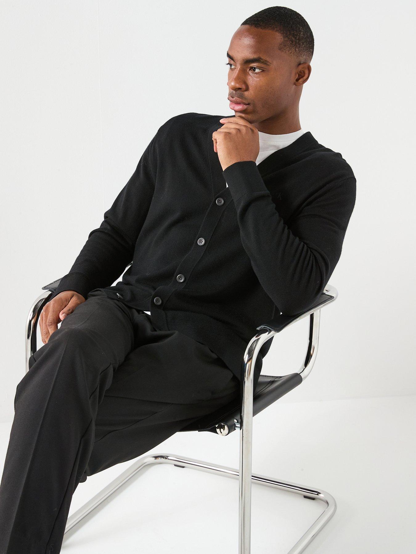 Male model poses on a shirt causally showing off his outfit that consists of a black cardigan, white t-shirt and black trousers. The perfect smart causal outfit for men.