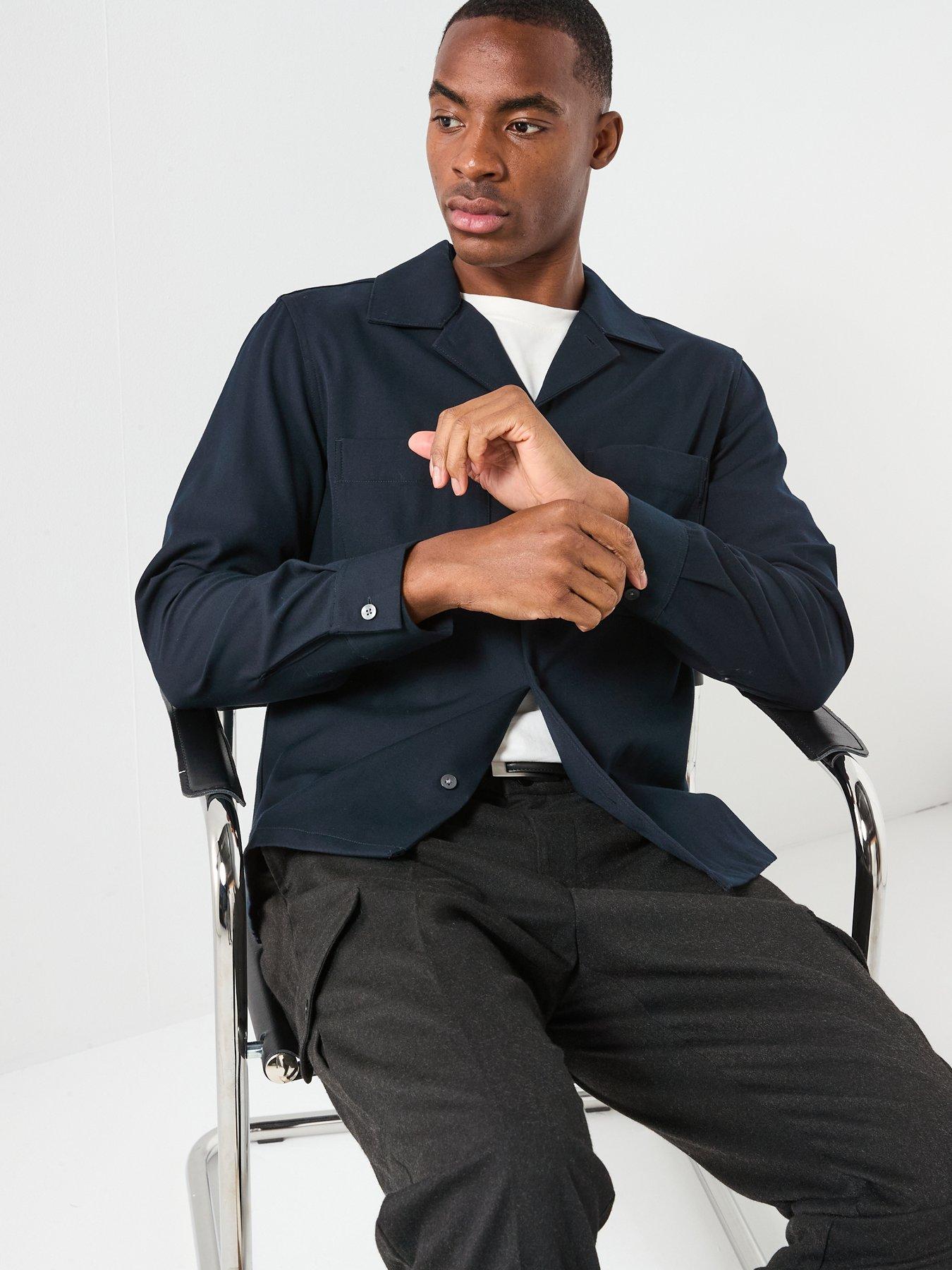 Man poses while siting on chair showing off his smart casual outfit of a white t-shirt and overshirt. 
