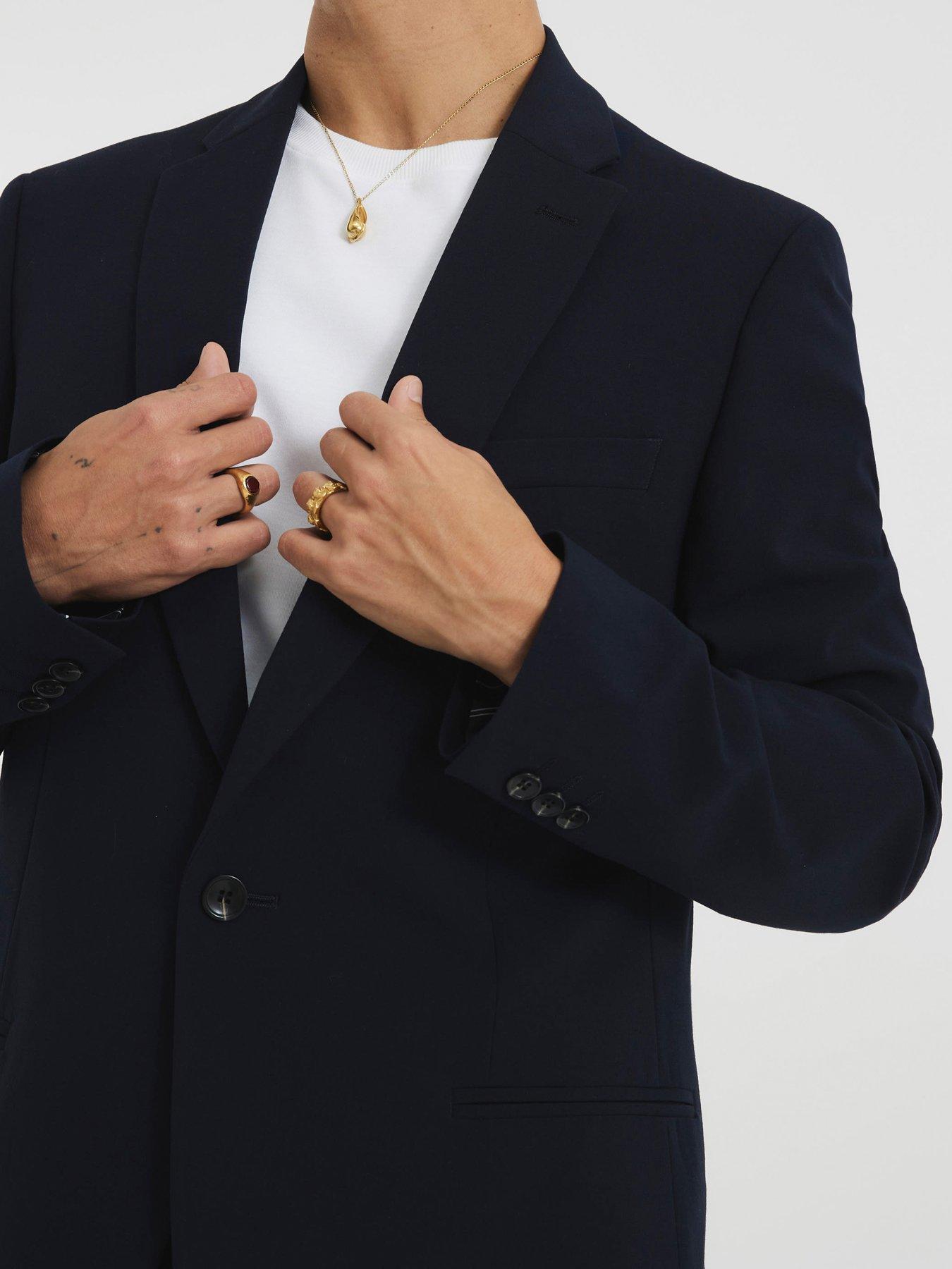 Close up of a man wearing a white t-shirt with an oversized black blazer over the top. The men's smart casual outfit has been accessoried with some men's jewellery like gold rings and a necklace.