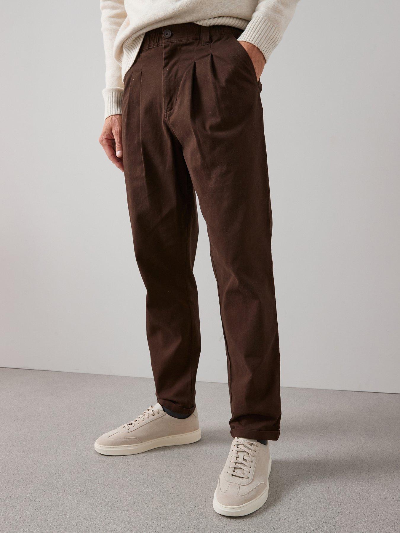Man wearing brown casual trousers with pleated detail at the waistband. The man has paired these trousers with a pair of causal white trainers.