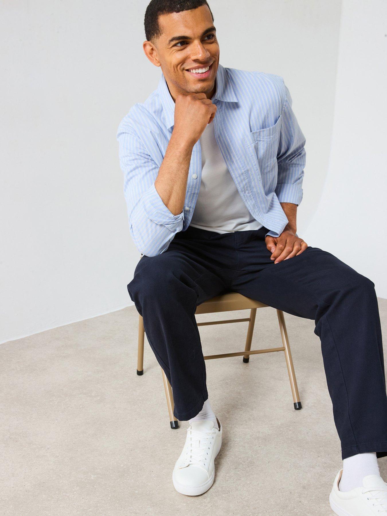 A man poses while sitting on a stool showing off his smart casual outfit. The look consists of a light blue striped shirt over a white tee paired with navy chinos and white trainers.