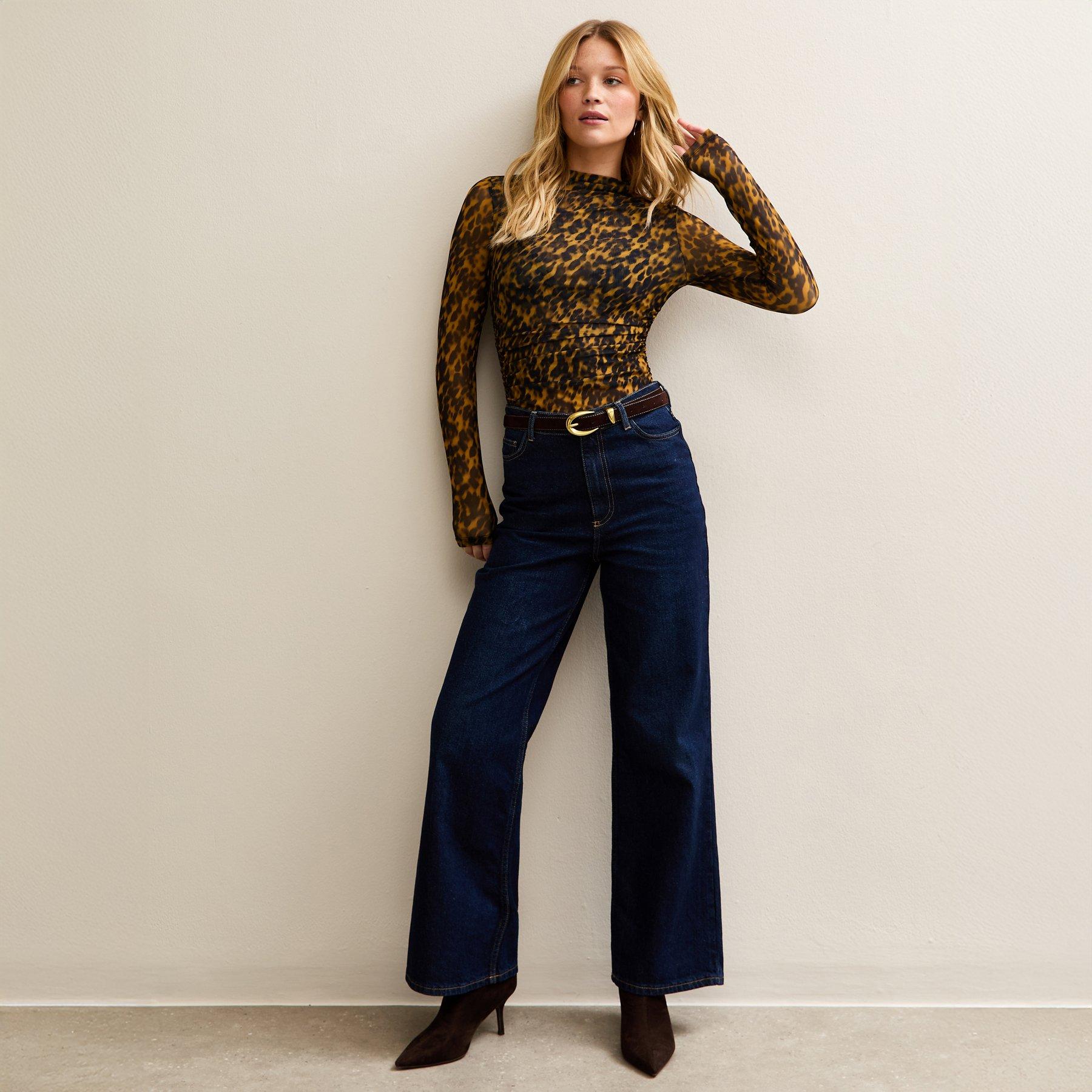 Woman poses wearing a long sleeve leopard print bodysuit tucked into a pair of indigo wide leg jeans. The outfit is paired with some pointy brown boots.