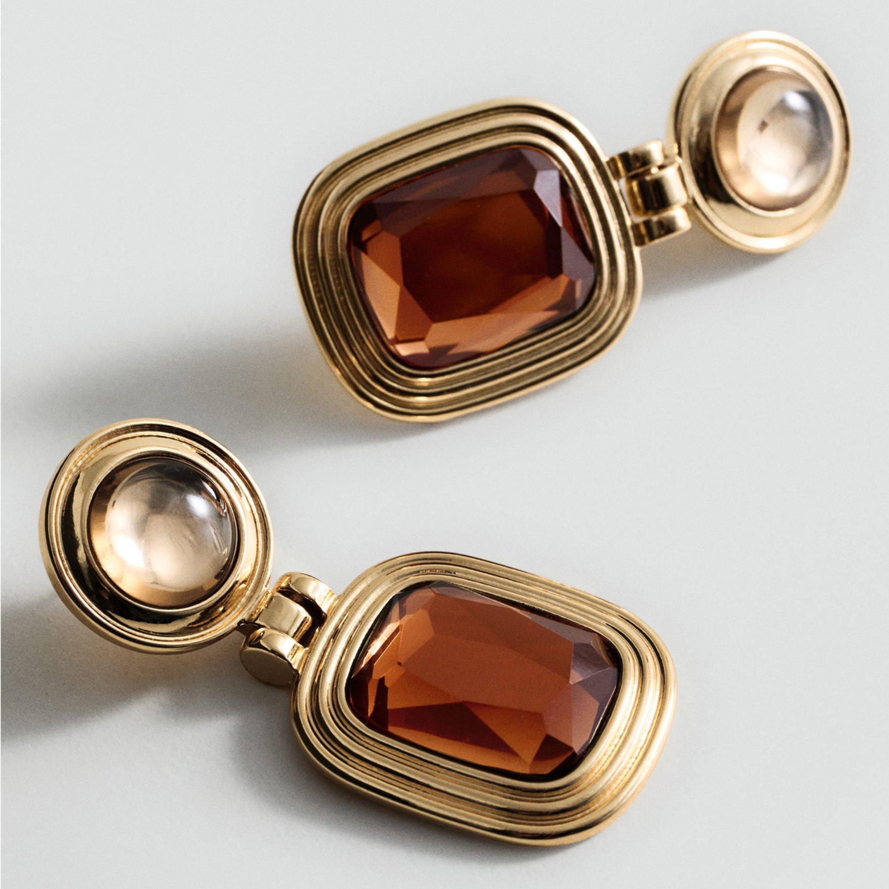 A pair of gold dangly earrings laying on a plain surface. The earrings have a large brown stone embellishment on the front.