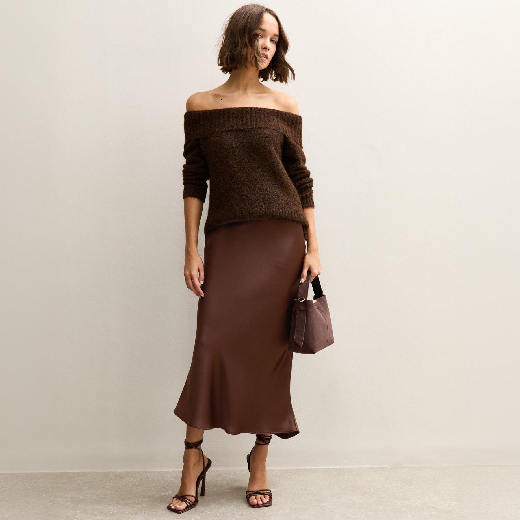 Woman poses wearing a classy date night outfit consisting of a bardot style brown jumper tucked into a similar toned satin midi skirt. The outfit is paired with a pair of heeled sandals and matching bag.