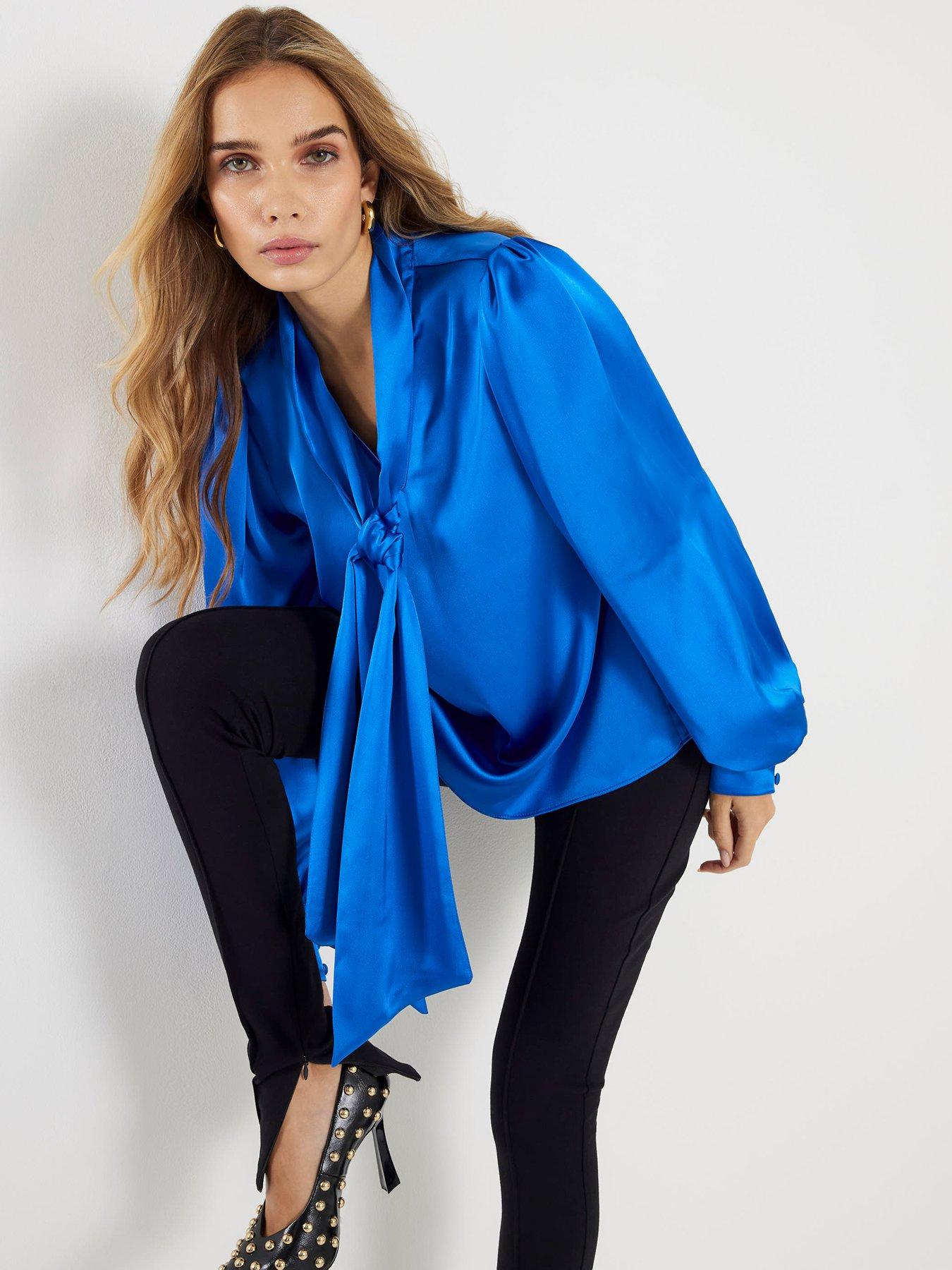 Model poses showing off her classy dinner date outfit. The outfit consists off a blue satin blouse with tie neck detail, paired with a pair of black tight trousers and black heels.