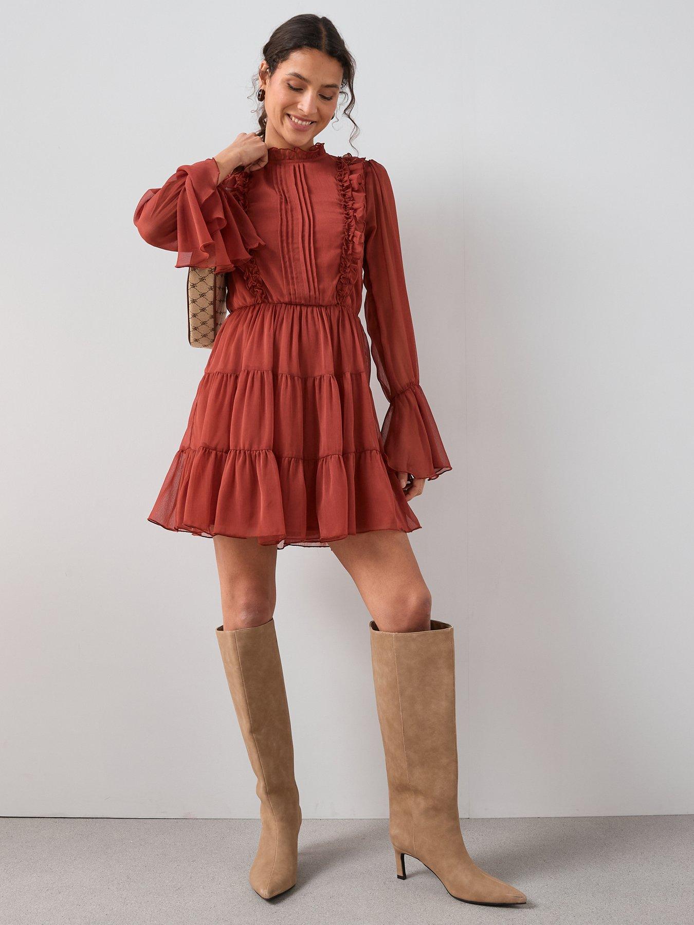 Model poses wearing a long sleeve floaty mini dress in a burnt orange colour. The boho outfit is paired with a beige pair of suede knee high boots. The perfect outfit for a date.
