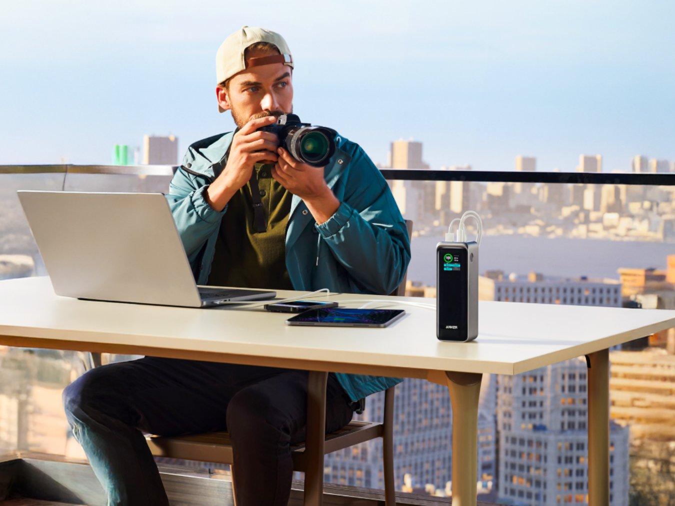 A man sits on a balcony with a city behind him and a power bank on a desk in front of him charging his laptop and tablet.